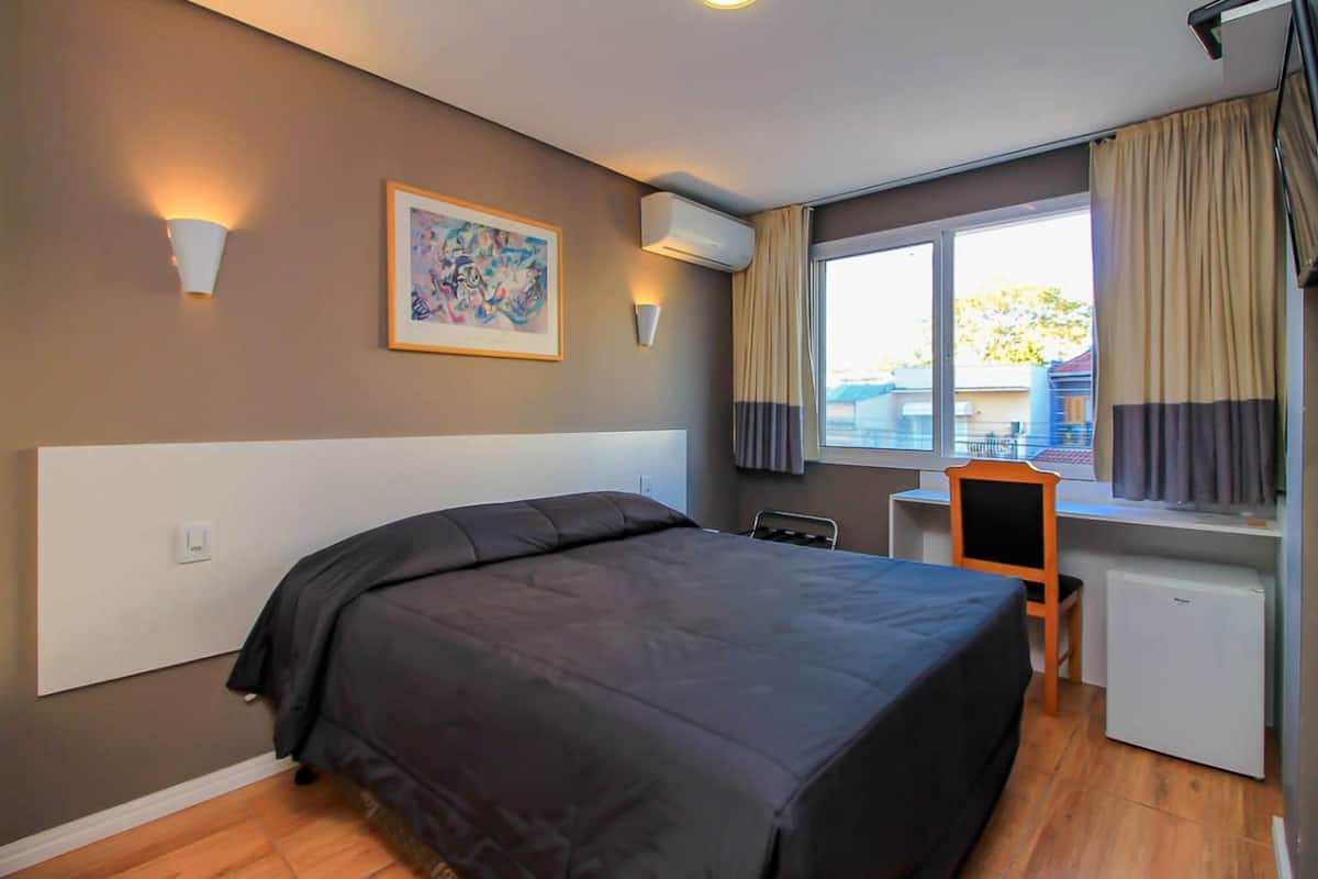 A hotel room with a bed, a desk, a chair, a small refrigerator, and a window with curtains.