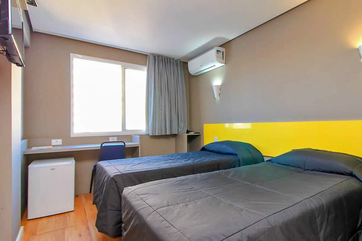 A compact hotel room with two beds, a small desk, a chair, a refrigerator, and a window with curtains.