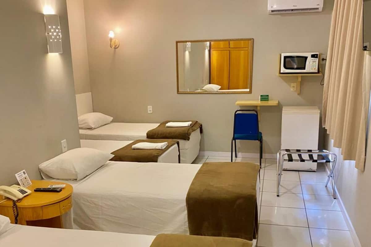 A hotel room with three beds, a desk, a chair, a microwave, and an air conditioning unit.