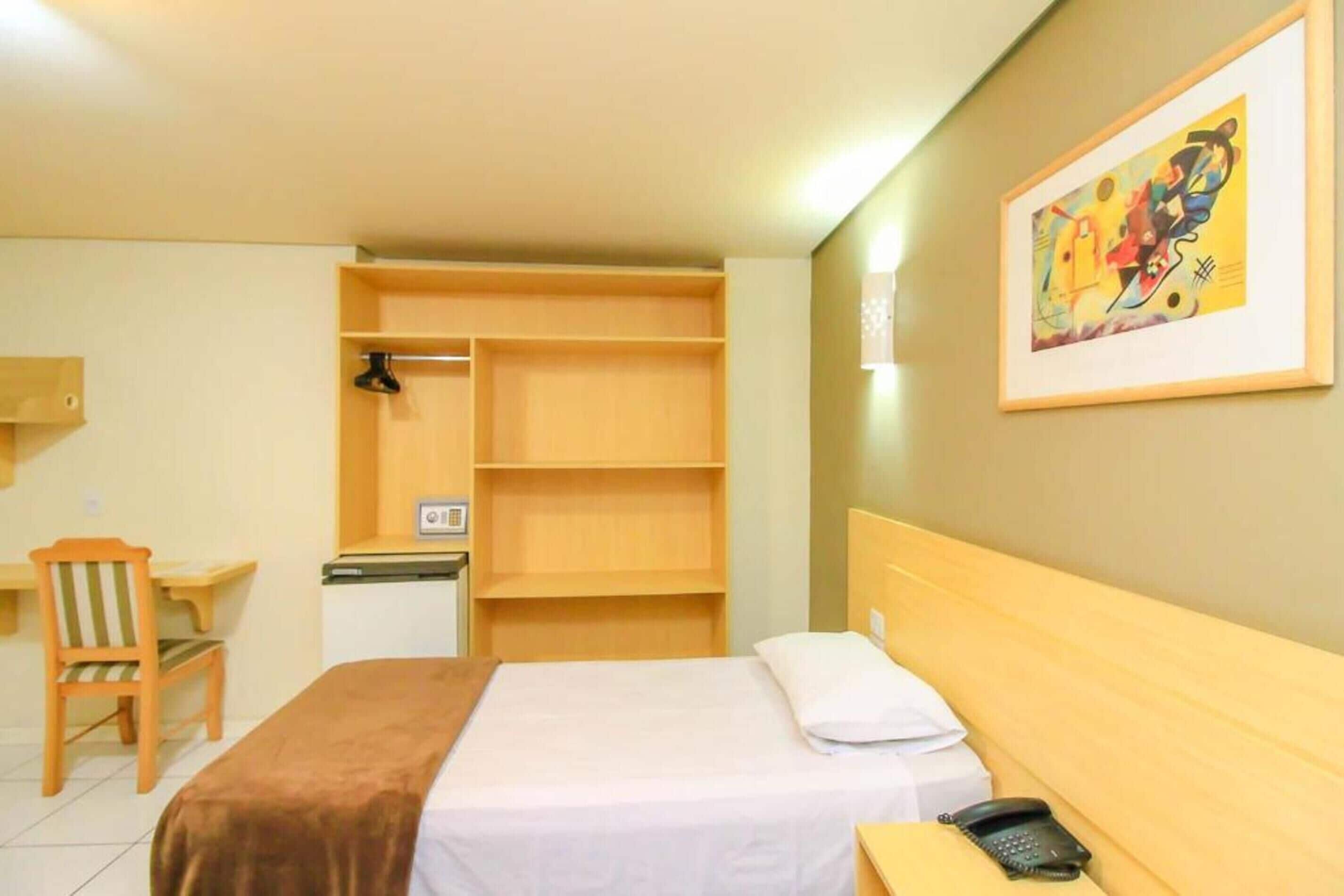 A hotel room with a bed, a desk, a chair, a wardrobe, and a painting on the wall.