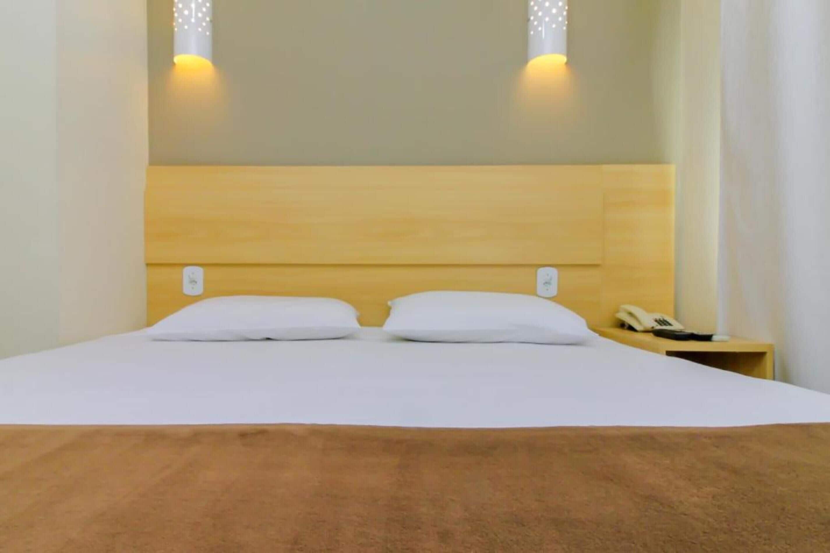 Una cama bien tendida con ropa de cama blanca y dos almohadas, un cabecero de madera y una mesita de noche con un teléfono.