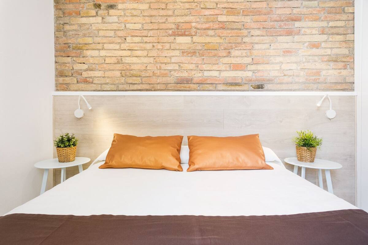 Een bed met twee kussens, een houten hoofdbord, twee kleine ronde tafels met plantenpotten, en een bakstenen muur erboven.