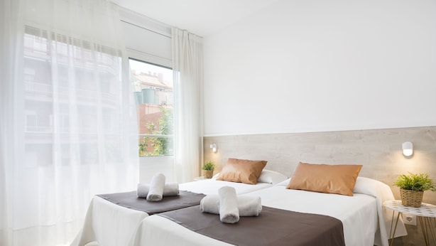 Een hotelkamer met twee bedden, een groot raam met dunne gordijnen, een klein bijzettafeltje met een plant en een wandlamp.