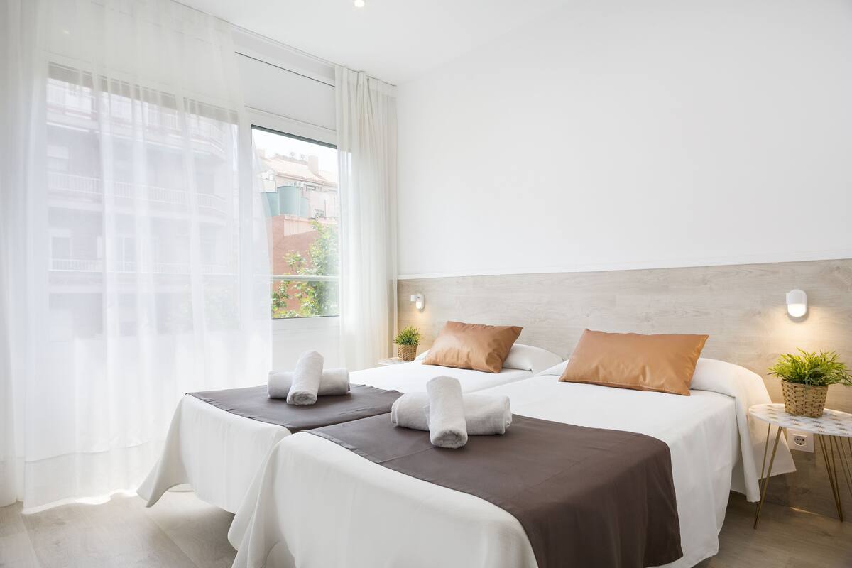 Een hotelkamer met twee bedden, een groot raam met dunne gordijnen, een klein bijzettafeltje met een plant en een wandlamp.