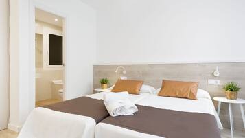 Habitación de hotel con dos camas, cabecero de madera, paredes blancas, una mesita de noche blanca con una planta y un baño visible a través de una puerta abierta.