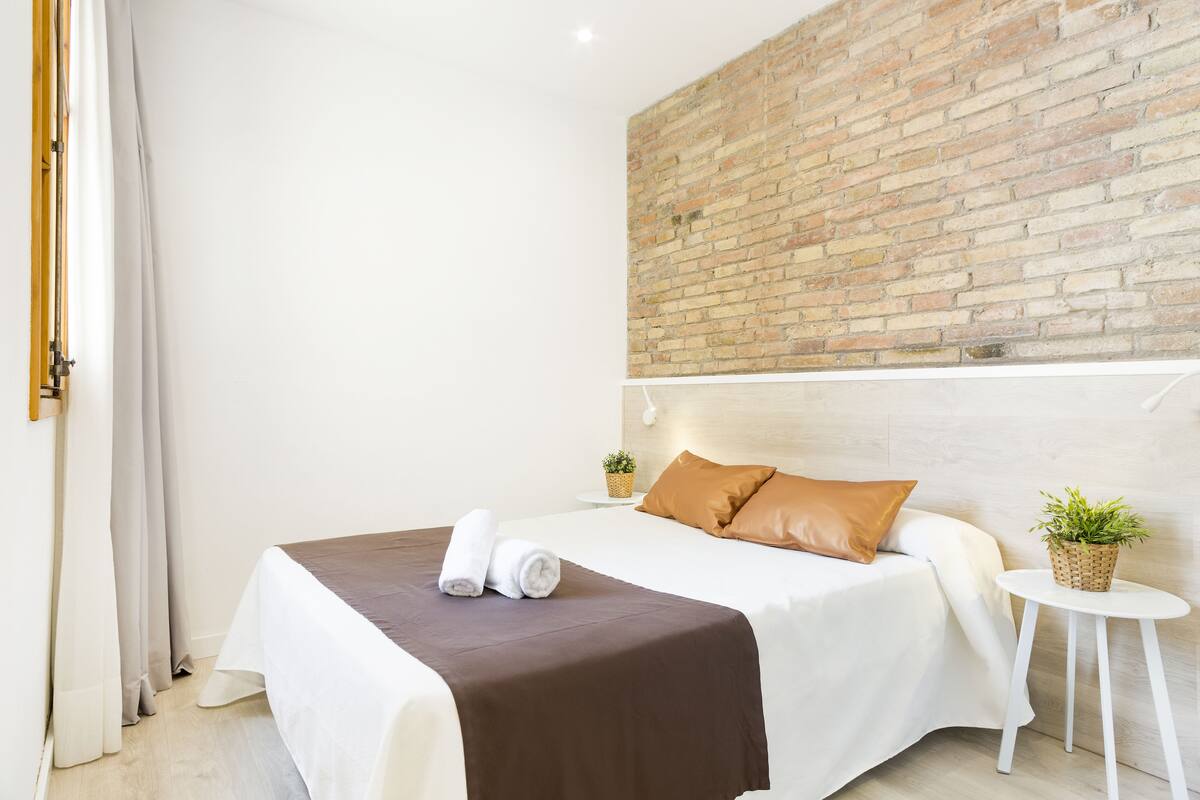 Een hotelkamer met een bakstenen muur, een bed met wit en bruin beddengoed, een klein rond tafeltje met een plant, en een wit gordijn.