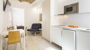 Een moderne keuken met witte kasten, een magnetron en een kleine koelkast. Er is een zithoek met een tafel en stoelen, en een stoel met een plaid.