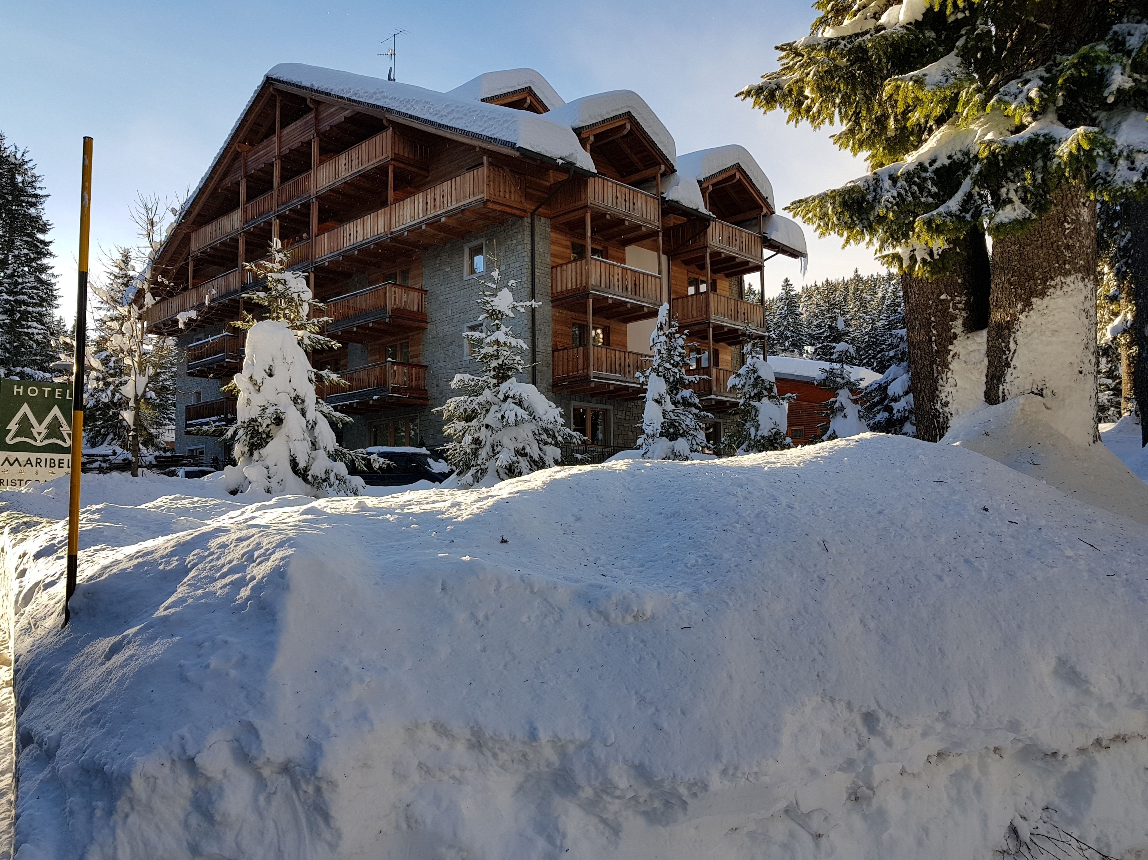 A multi-story wooden building with balconies, surrounded by snow-covered trees and a sign indicating a hotel.