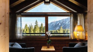 Una habitación acogedora con techo de madera, un ventanal con vista a la montaña, dos sofás oscuros, una mesita con una planta y una lámpara.