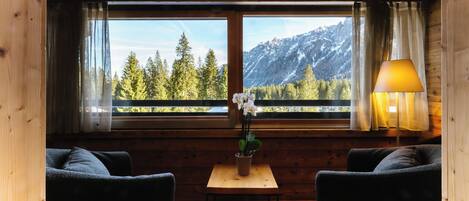 A cozy room with a wooden ceiling, a large window with a mountain view, two dark sofas, a small table with a plant, and a lamp.
