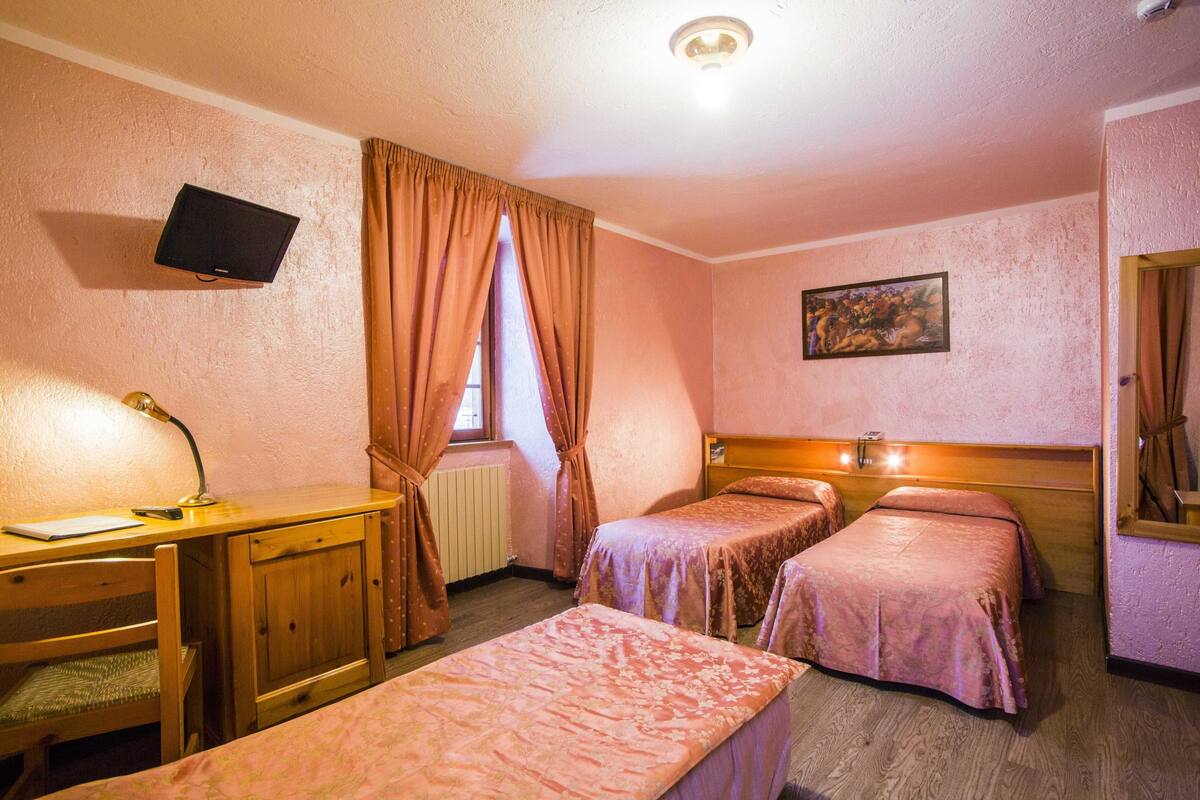 A room with two single beds, a desk with a chair, a television mounted on the wall, and a window with curtains.