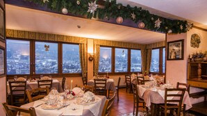 Un comedor con varias mesas dispuestas para una comida, grandes ventanales con vista a la ciudad y decoraciones festivas.