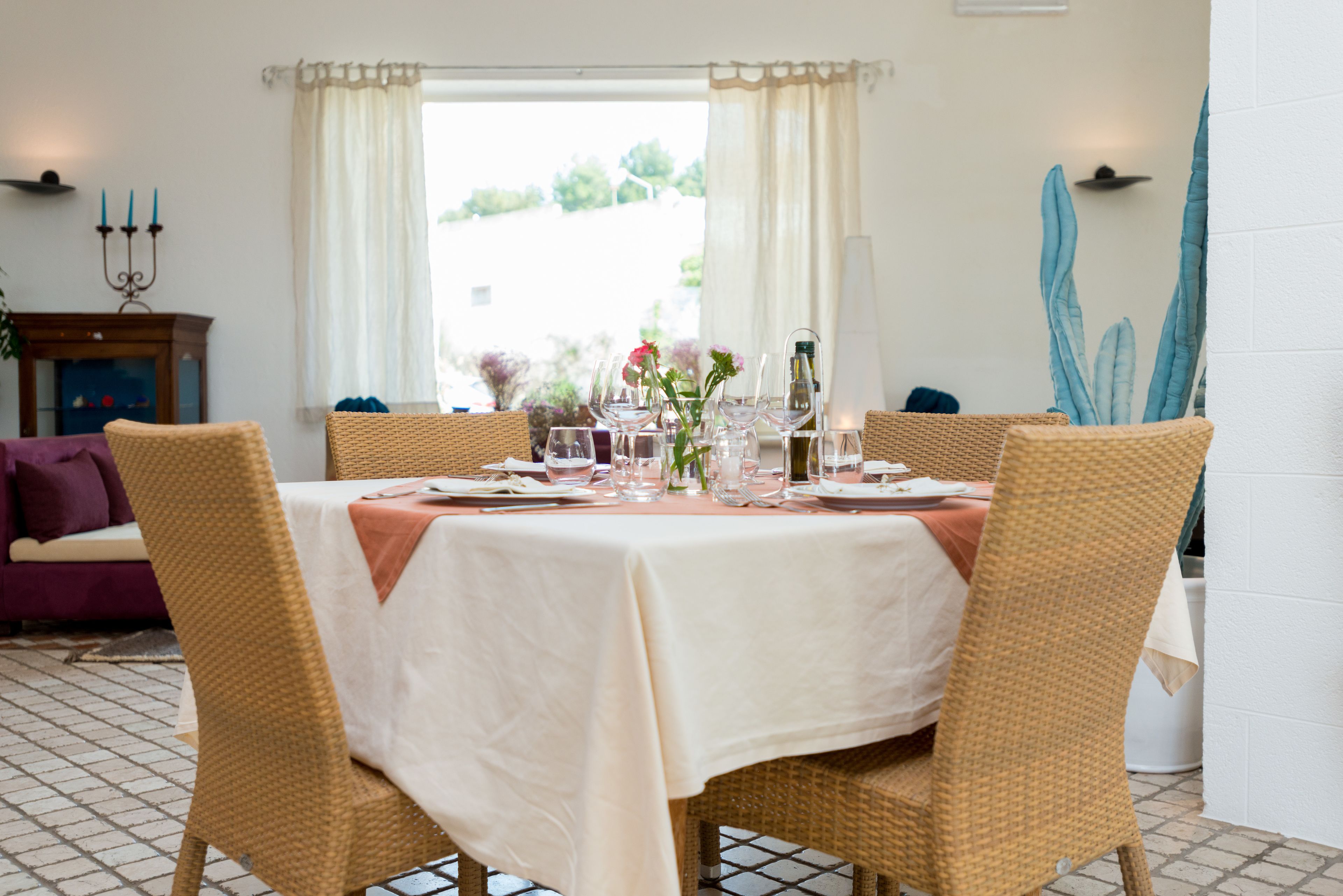 A dining area with a table set for four, surrounded by wicker chairs, a window with curtains, and a view of greenery outside.