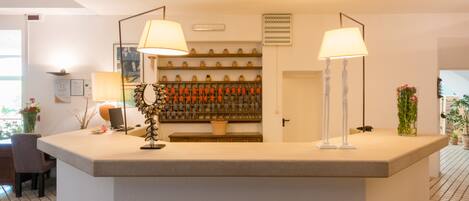 A reception area with a counter, two lamps, a wall-mounted shelf with bottles, a door, and a potted plant.