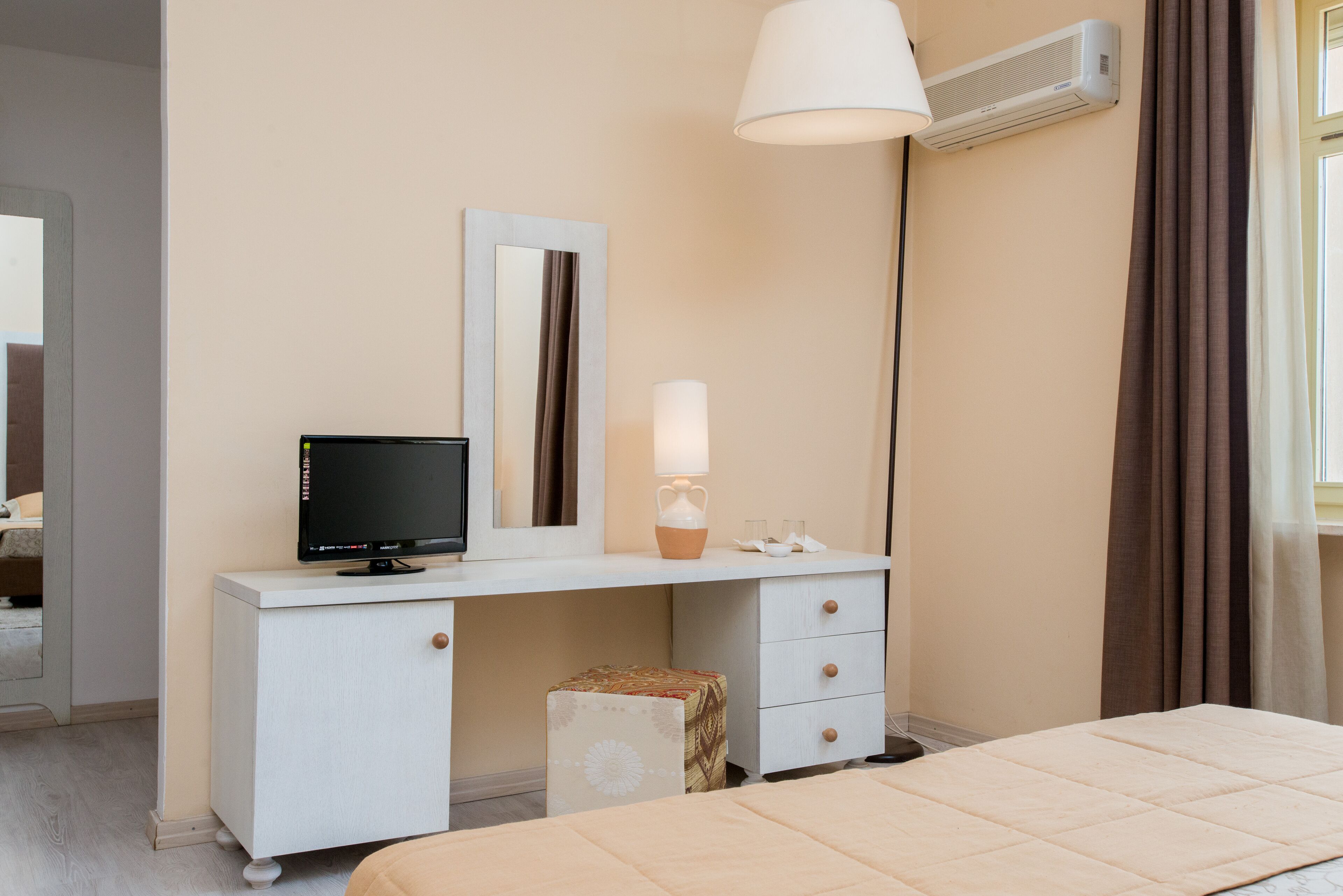 A hotel room with a bed, a desk with a computer, a mirror, a lamp, and an air conditioning unit.