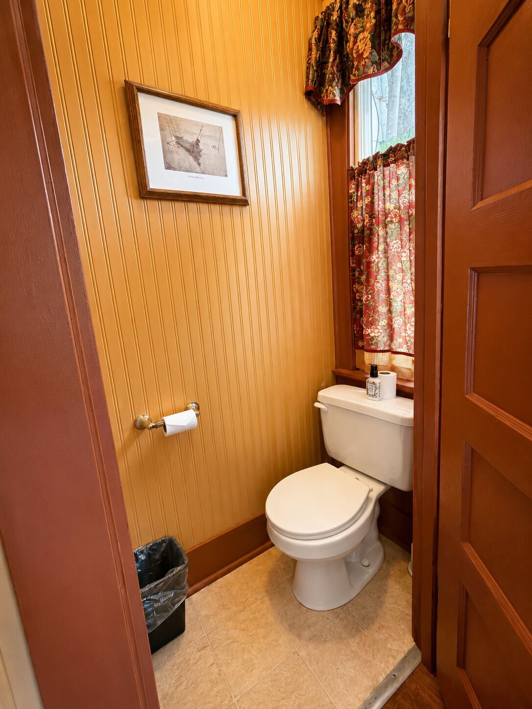 Une petite salle de bain avec des toilettes, un rouleau de papier toilette, une poubelle et un tableau encadré au mur.