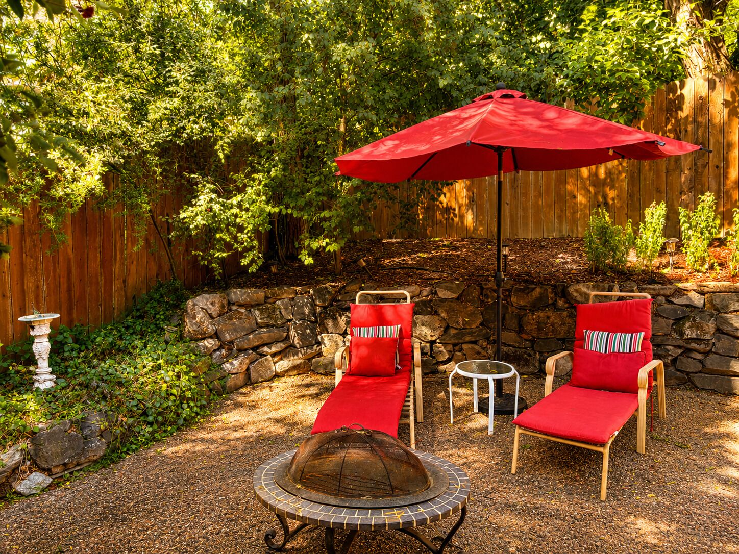 A patio with two red lounge chairs, a stone wall, a fire pit, and a red umbrella.