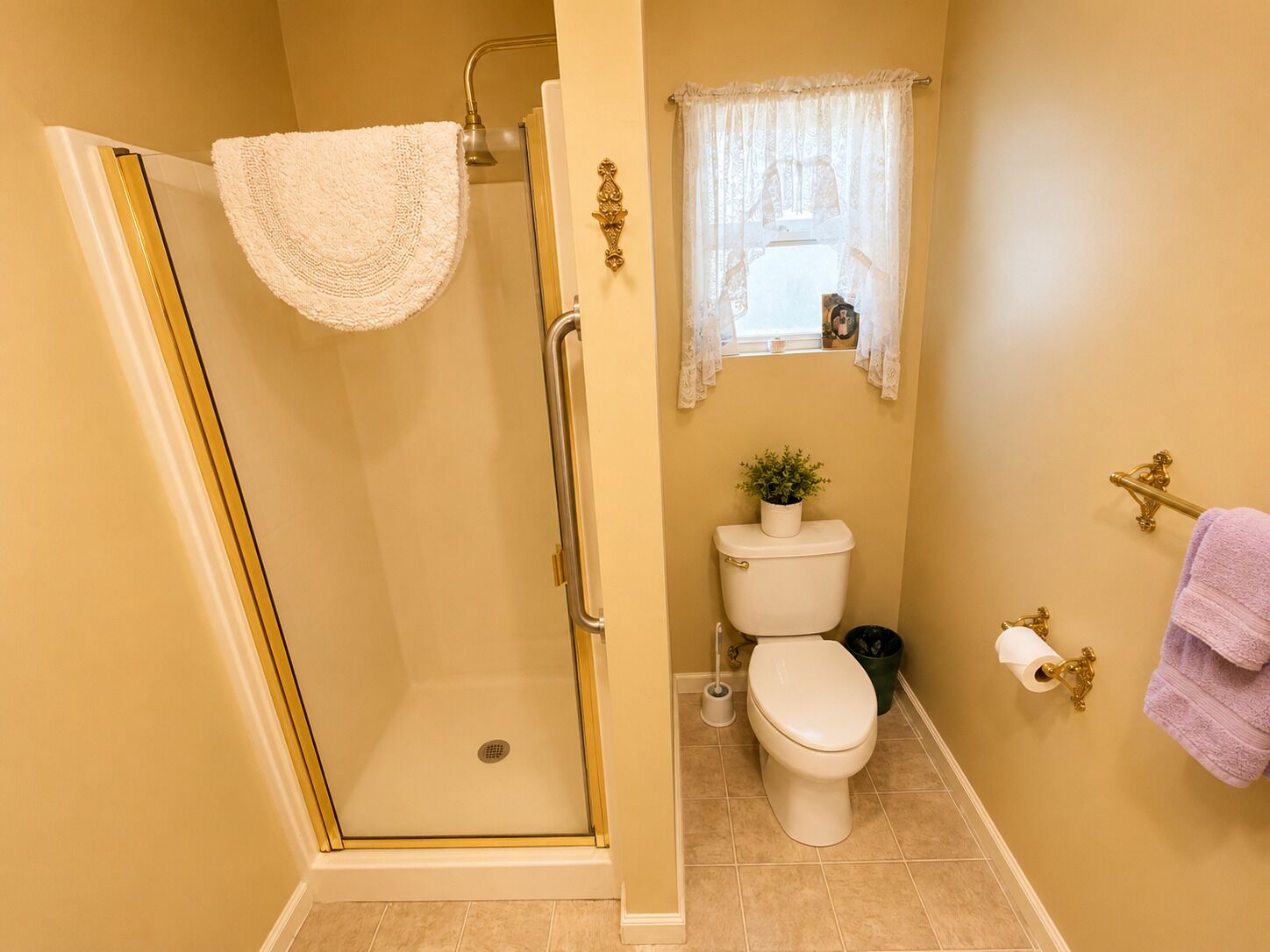 A bathroom with a glass-enclosed shower, a toilet, a small window with lace curtains, and a towel rack.