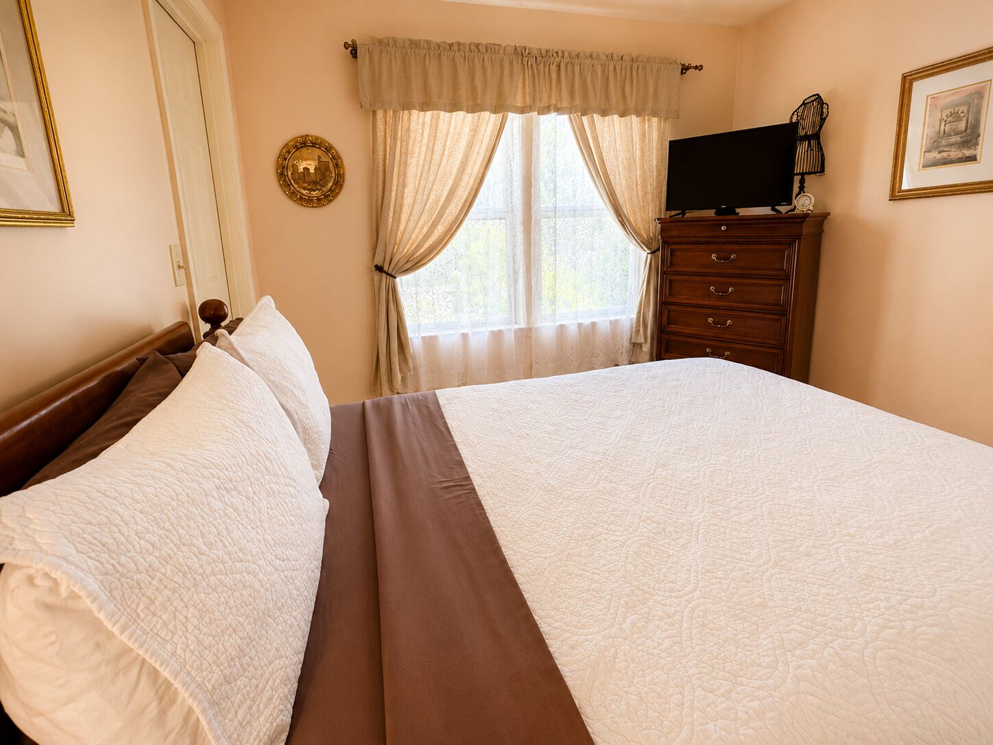 Una cama bien hecha con una colcha marrón y almohadas blancas, un tocador de madera con un televisor de pantalla plana y una ventana con cortinas traslúcidas.