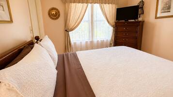 Una cama bien hecha con una colcha marrón y almohadas blancas, un tocador de madera con un televisor de pantalla plana y una ventana con cortinas traslúcidas.