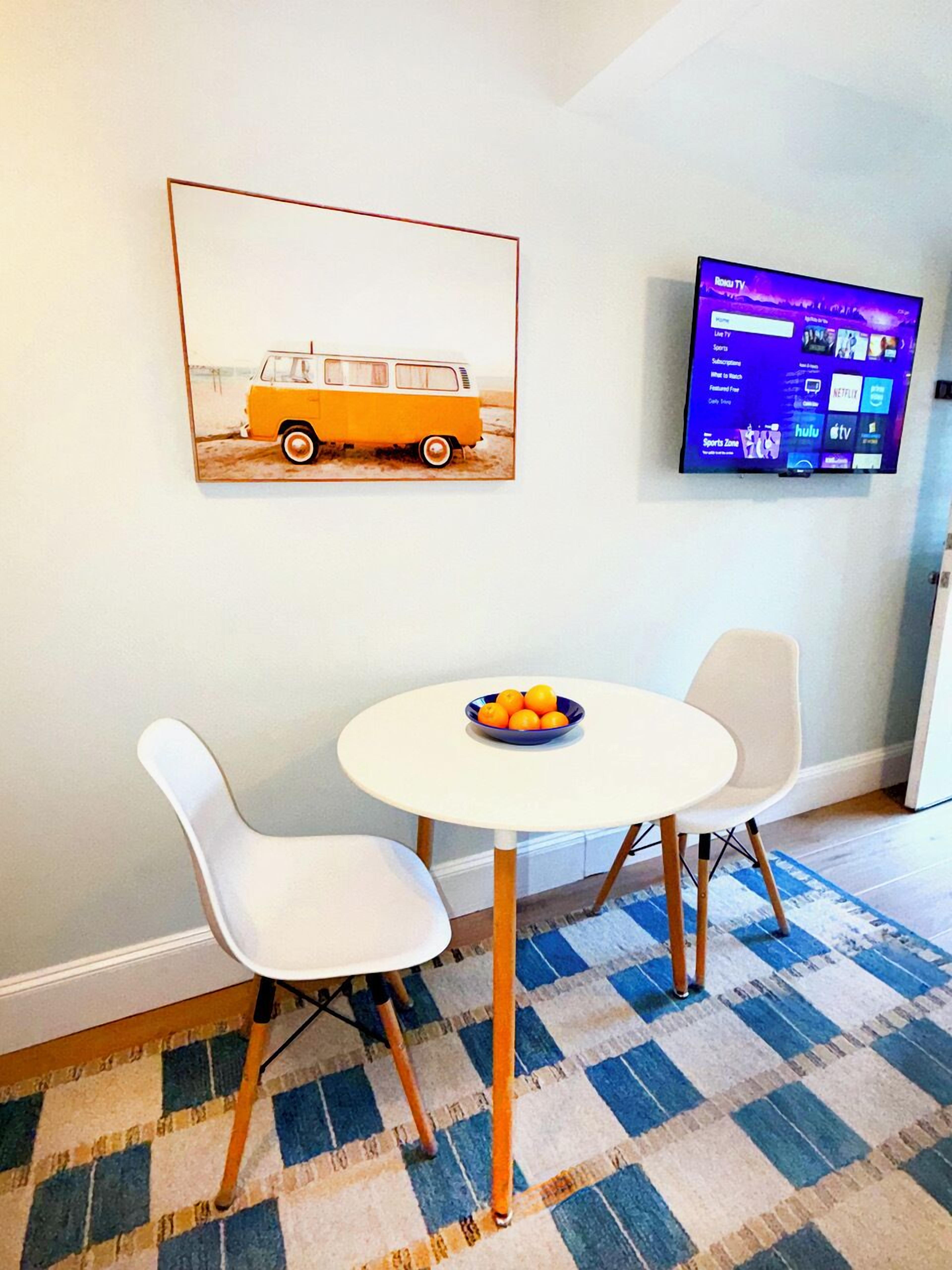 A room with a round table, two white chairs, a bowl of oranges, a framed picture of a van, and a mounted TV displaying a menu.