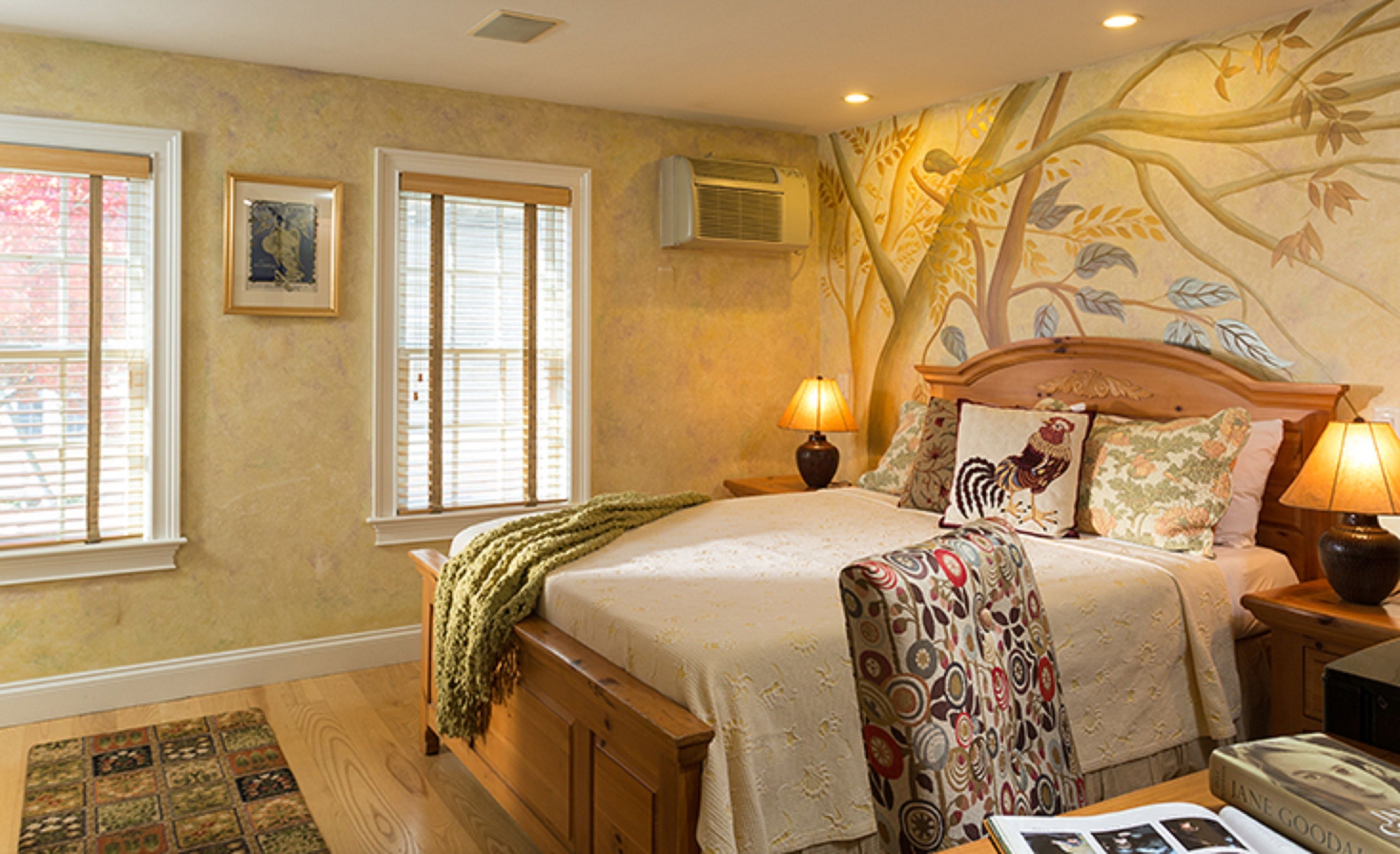 A bedroom with a large bed, two bedside lamps, a window, an air conditioner, and a patterned wall mural.