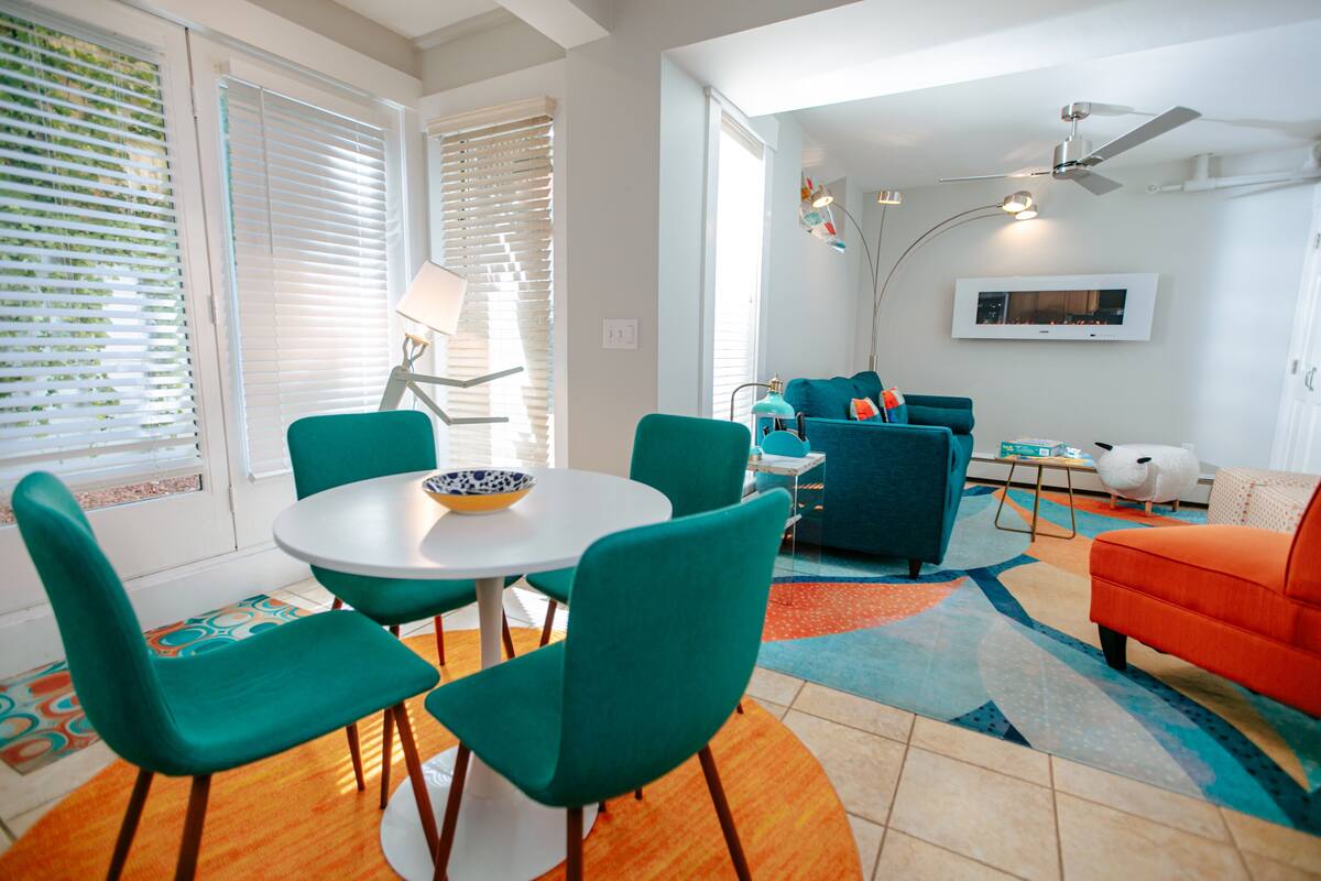 Una sala de estar moderna con una mesa redonda, sillas color turquesa, un sofá azul y un sillón naranja.
