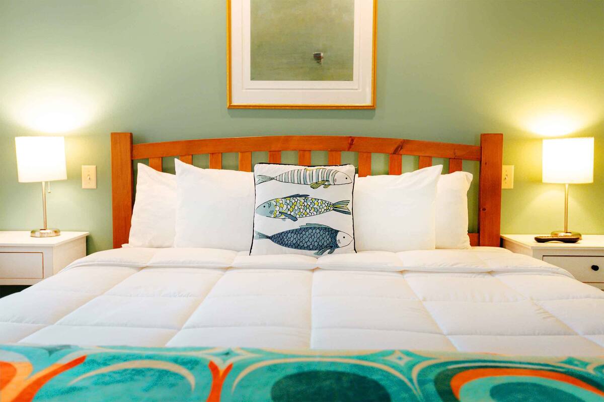 Una cama bien tendida con sábanas blancas y una almohada decorativa con motivos de peces. Hay dos lámparas de noche a cada lado de la cama.