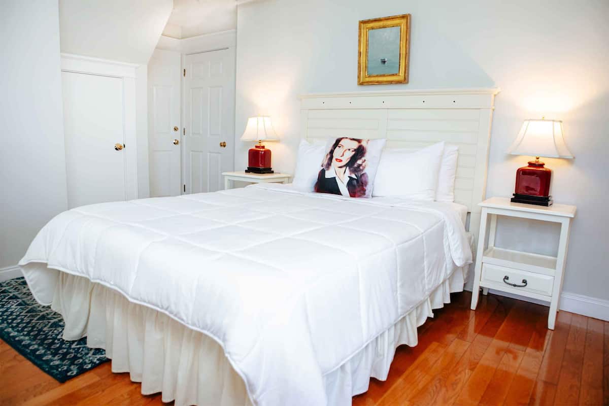 Un dormitorio ordenado con una cama grande, cabecera blanca, dos mesitas de noche con lámparas y una alfombra estampada.