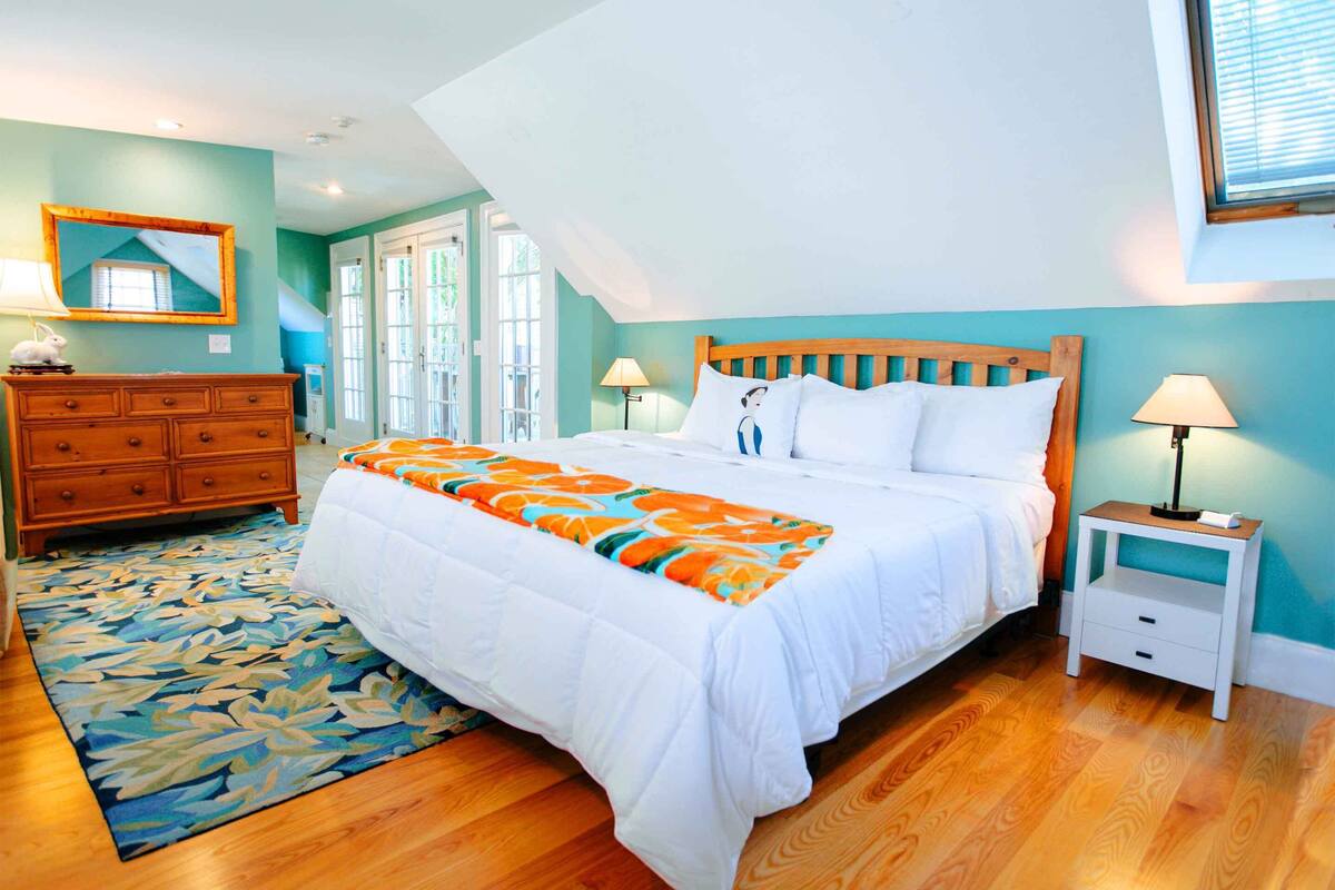 Un dormitorio con cama de madera, ropa de cama blanca, alfombra floral, cómoda de madera y mesita de noche con lámpara.