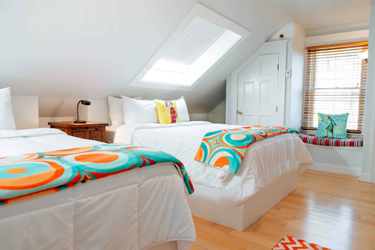 Un dormitorio con dos camas, un tragaluz, una ventana con persianas y una mesita de noche con lámpara.