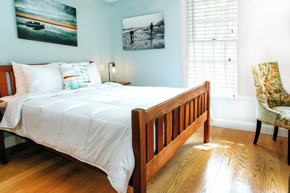 Un dormitorio con una cama de madera, ropa de cama blanca, una mesita de noche, una lámpara y dos obras de arte en la pared.