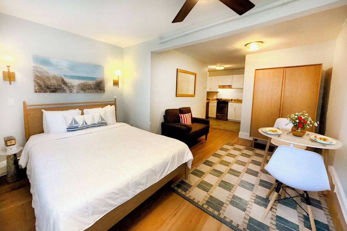 Una habitación de hotel con cama, una silla, una mesita con un jarrón y una zona de cocina al fondo.