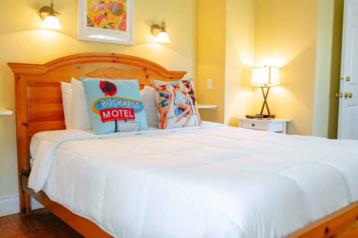 Una cama bien hecha con cabecera de madera, dos lámparas fijadas a la pared y un cuadro enmarcado sobre la cama.