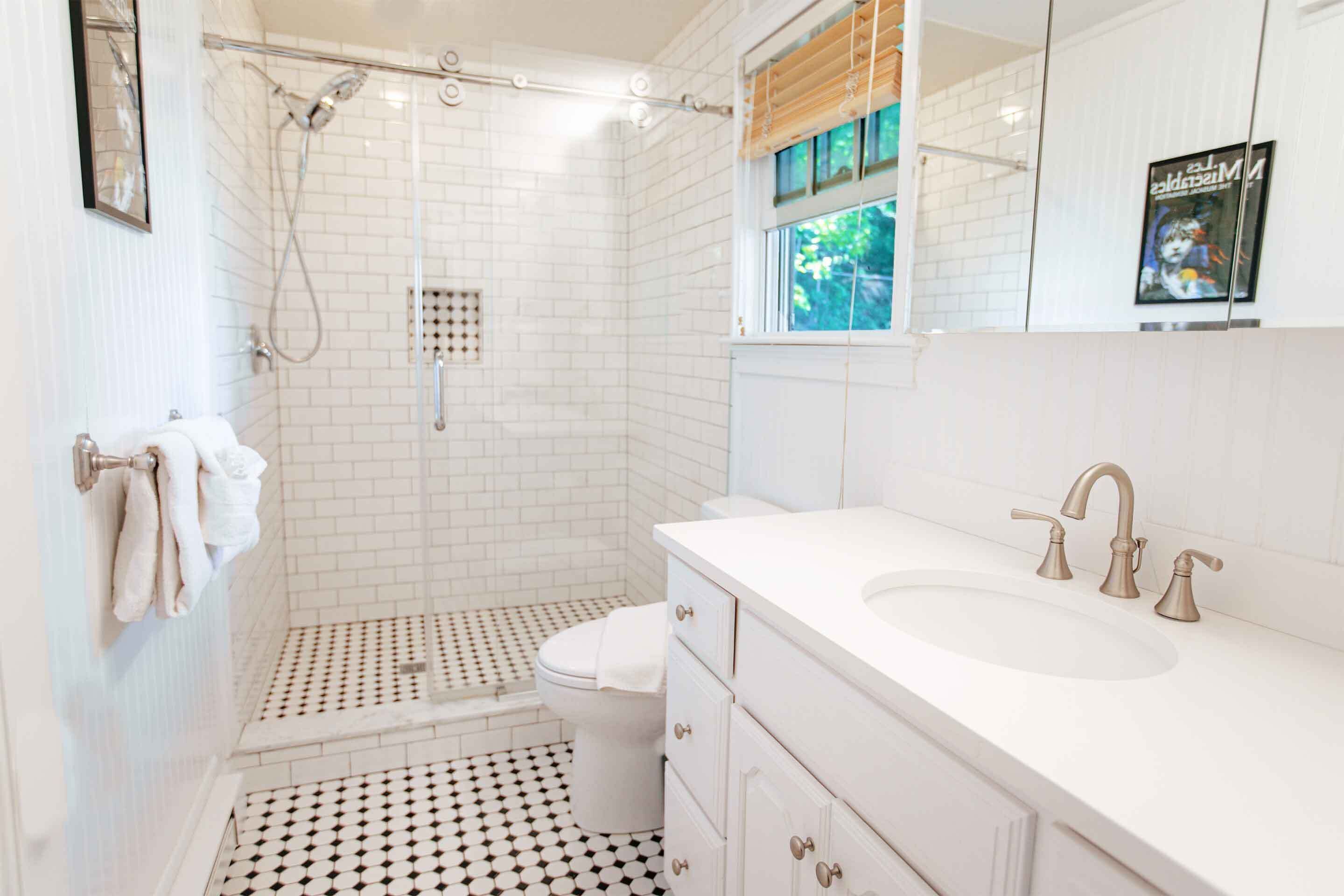 A bathroom with a white tiled shower, a toilet, a sink with a mirror, and a window with blinds.