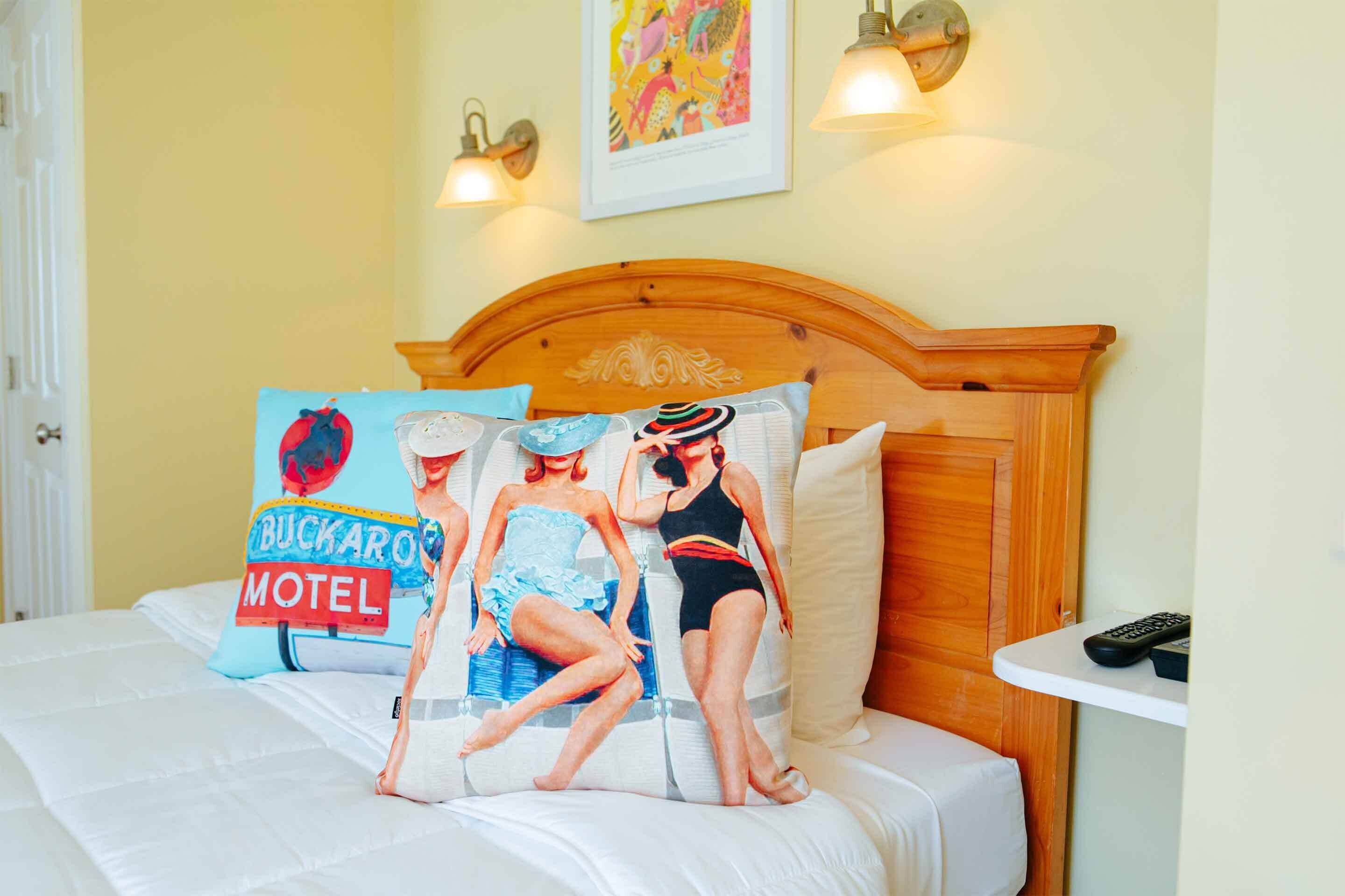 Uma cama com cabeceira de madeira, duas luminárias fixadas na parede e um travesseiro com a placa 'Buckaroo Motel'.
