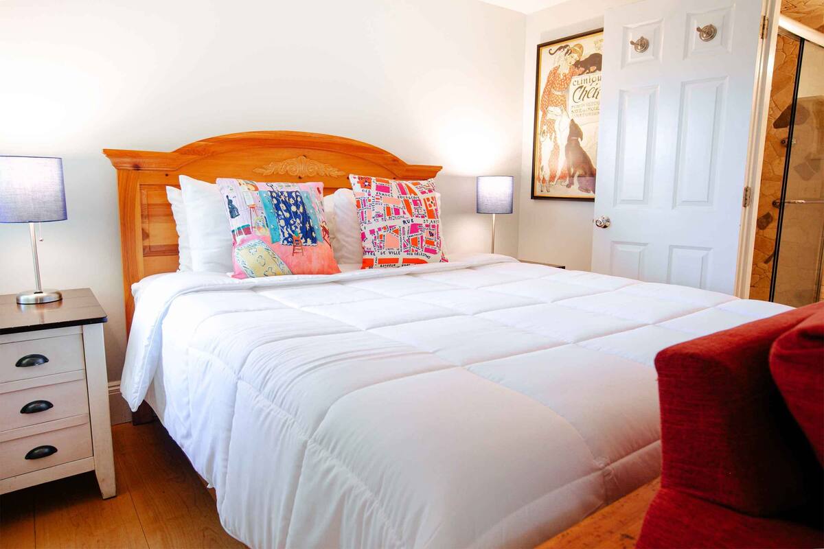 Una cama bien hecha con cabecera de madera, ropa de cama blanca y almohadas coloridas. Se ve un mesita de noche con una lámpara y una silla roja al frente.