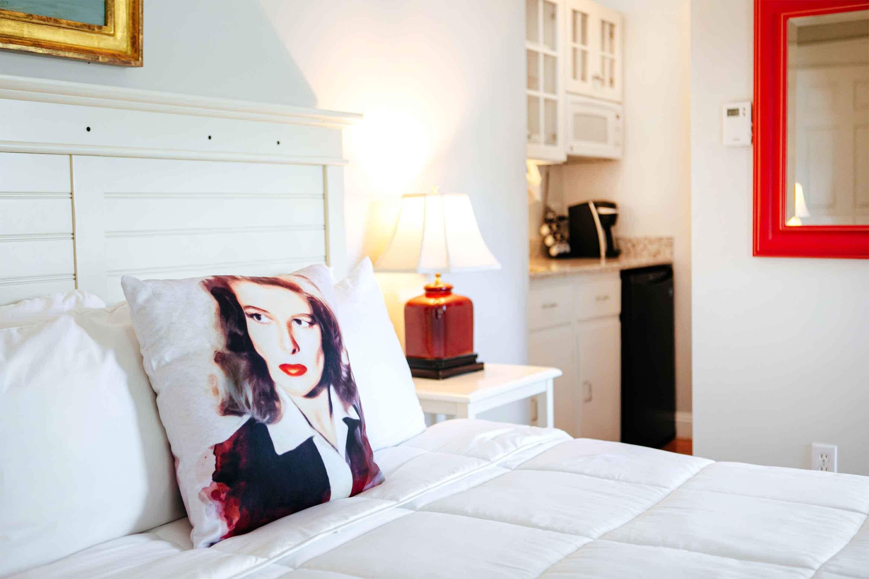 A hotel room with a bed, a pillow with a printed design, a bedside lamp, a white nightstand, and a red-framed mirror.