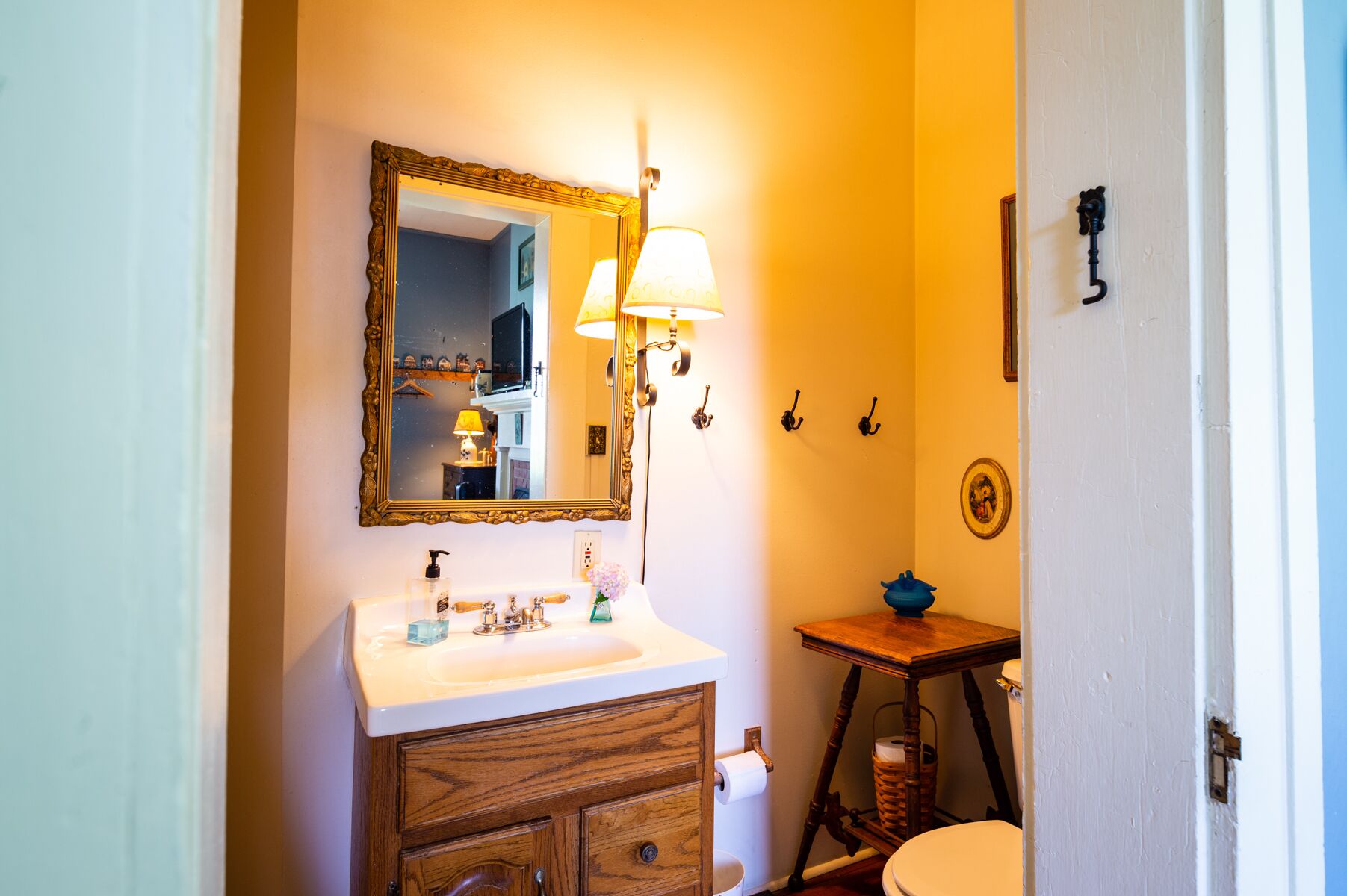 Une salle de bain avec un meuble-lavabo en bois, un lavabo blanc, un miroir doté d’un cadre orné, une lampe fixée au mur et une petite table en bois sur laquelle est posé un bol bleu.
