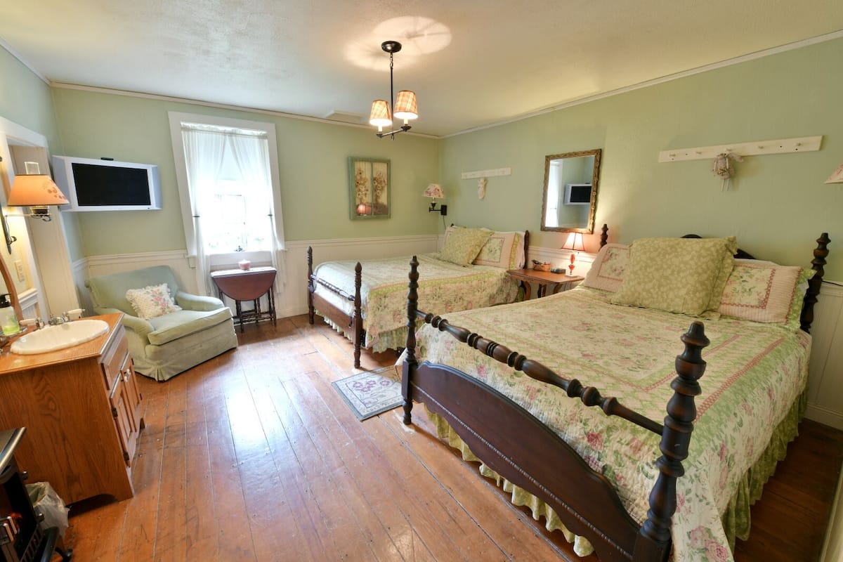 A bedroom with two single beds, a wooden dresser, a vanity with a mirror, a window with curtains, and a ceiling light fixture.