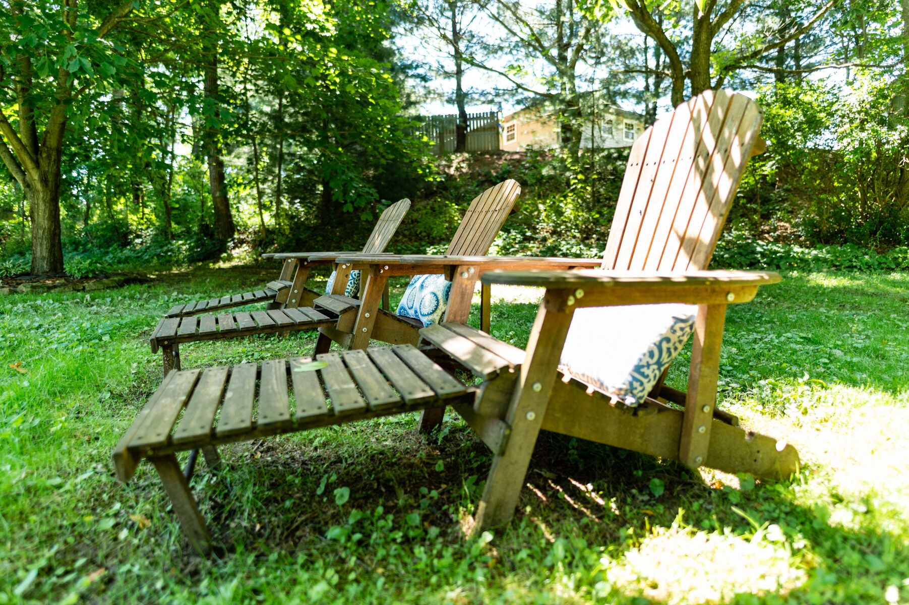 A set of wooden Adirondack chairs with cushions, positioned on a grassy area surrounded by trees.