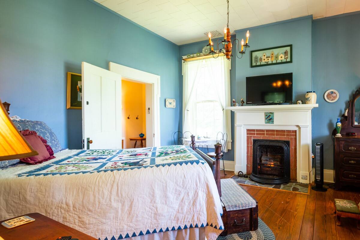A bedroom with a large bed, a fireplace, a television, and a door leading to another room.