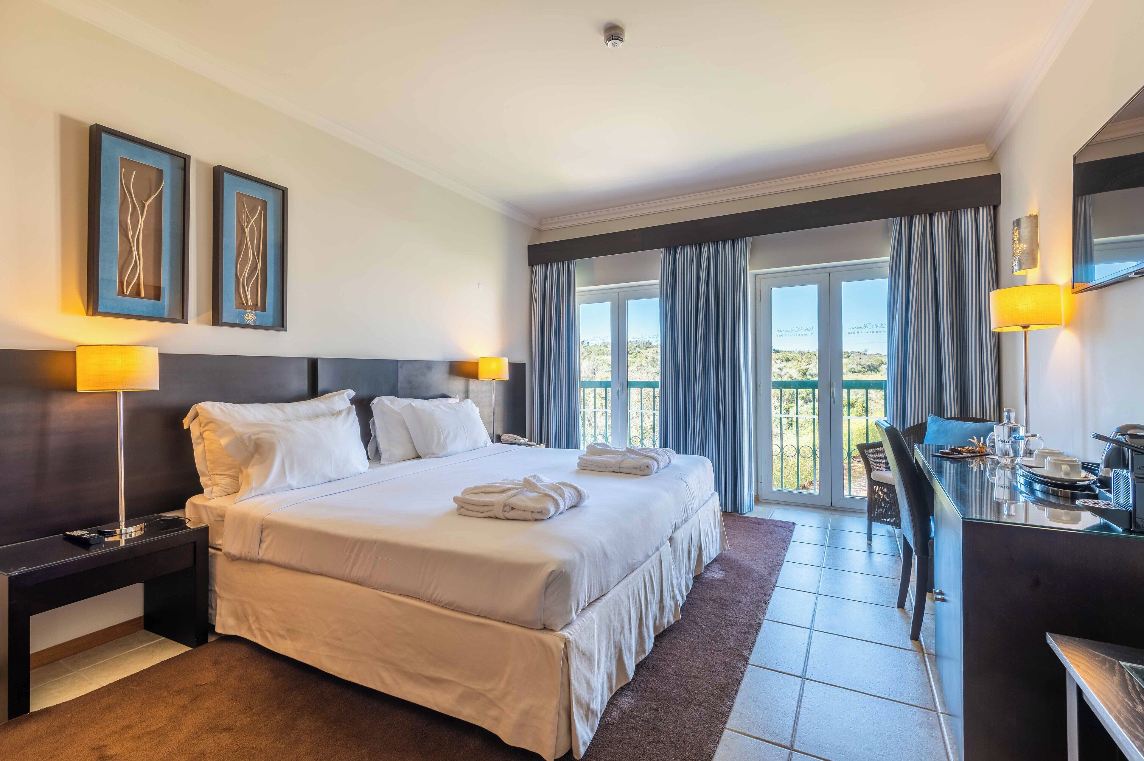 A hotel room with a large bed, two bedside lamps, a desk with a chair, and a view of the outdoors through a sliding door.