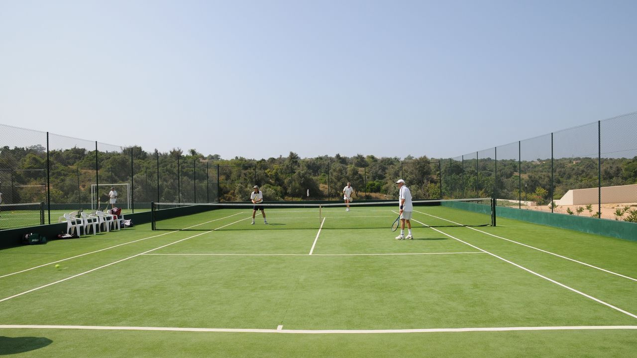 Una cancha de tenis al aire libre con superficie verde, rodeada por una valla negra y árboles al fondo.