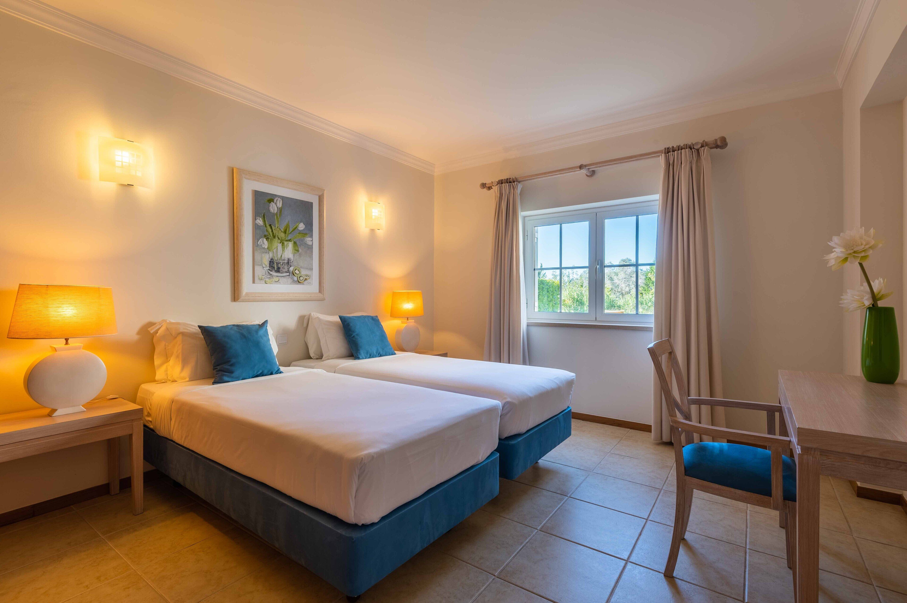 A hotel room with two beds, a dining table, a chair, and a window with curtains.
