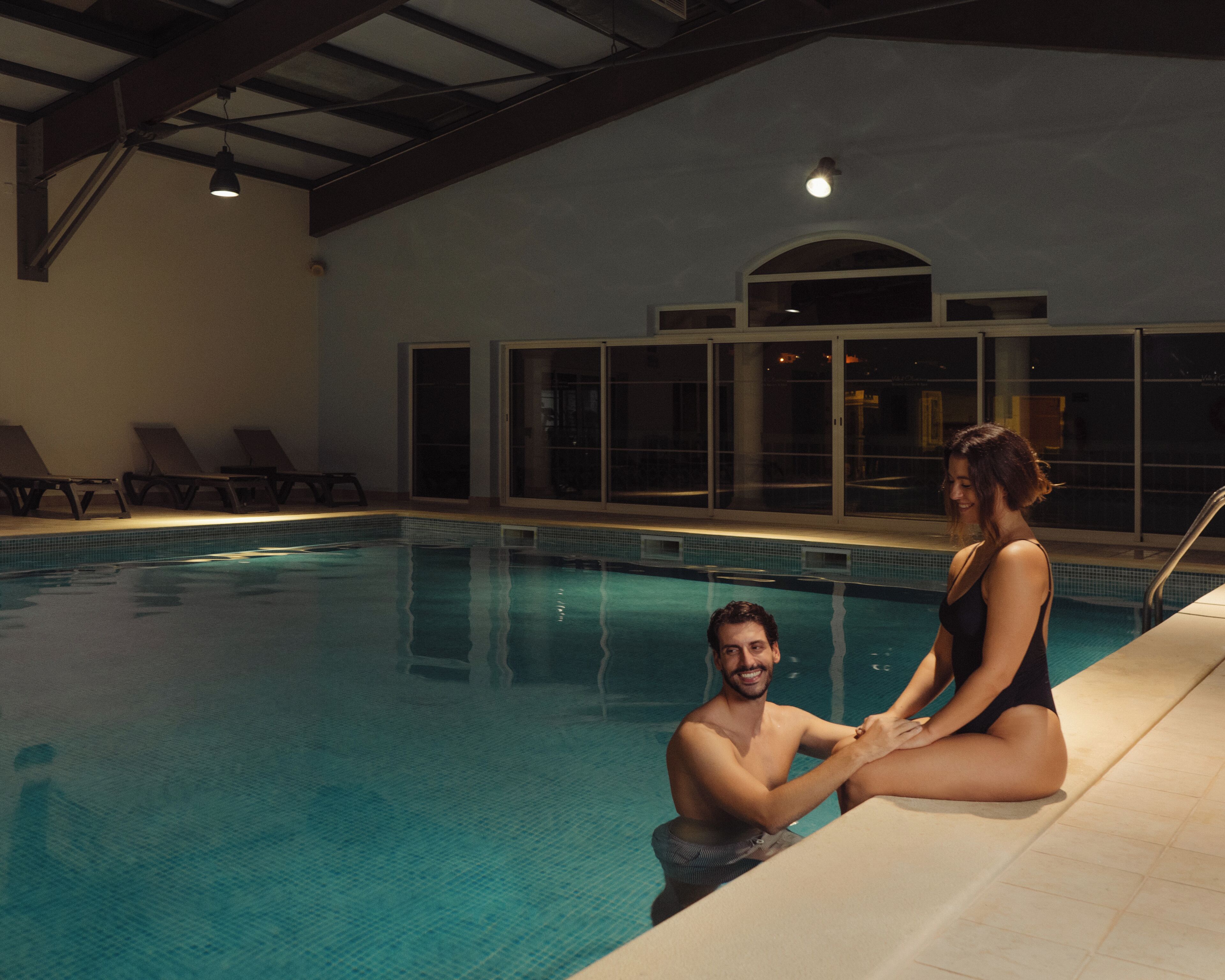 Una piscina interior con agua cristalina, una pareja sentada al borde, sillas de descanso y un amplio ventanal de arco.
