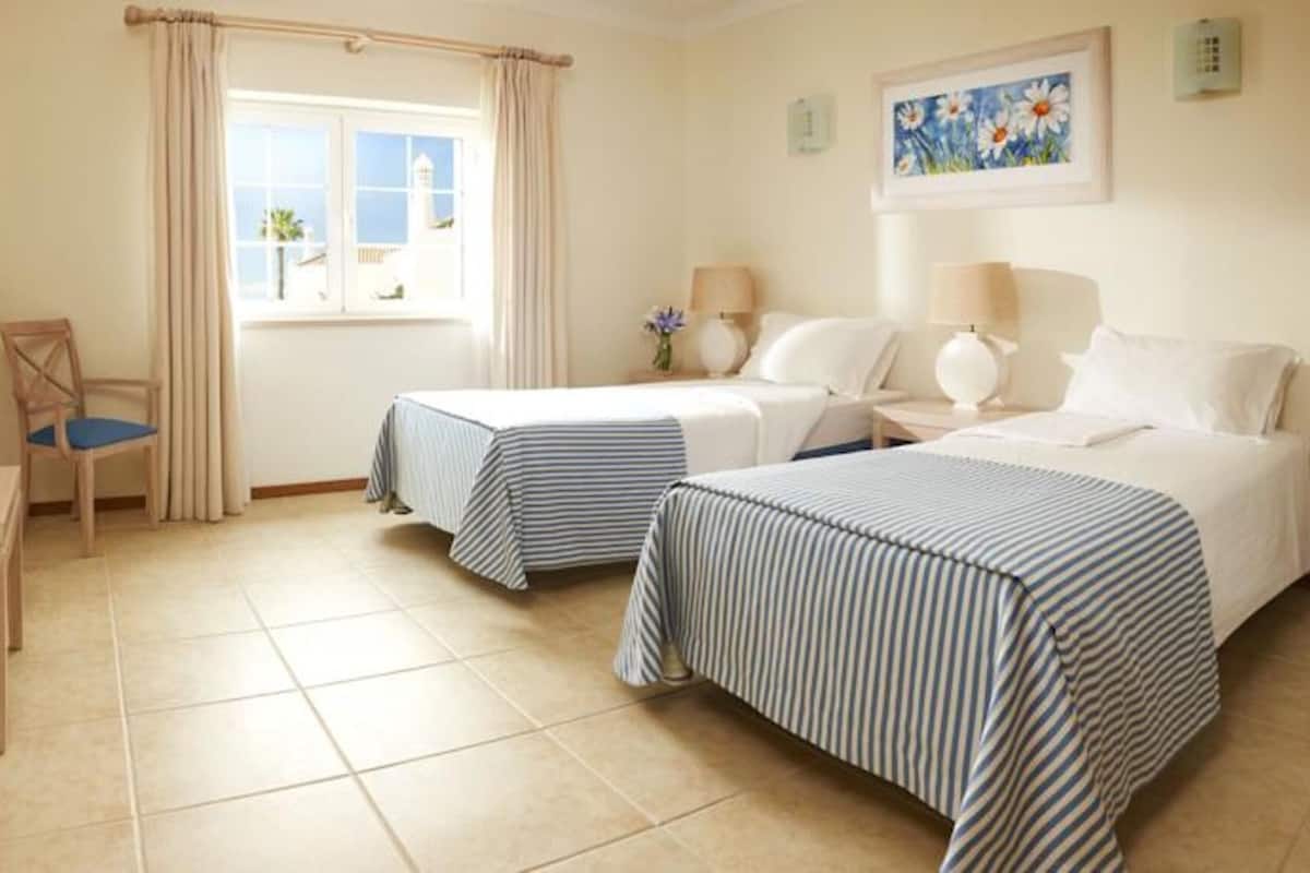 A hotel room with two beds, a chair, a small table, and a window with curtains.