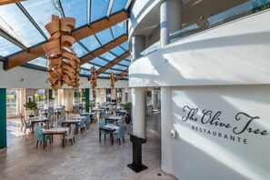 Un interior de restaurante con diseño moderno, que cuenta con techo de vidrio, vigas de madera y un letrero que dice 'The Olive Tree Restaurantante'.