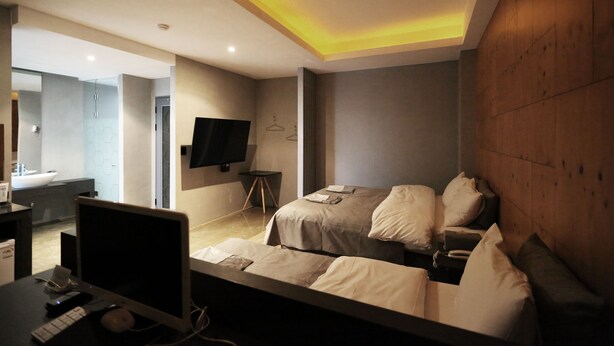 Une chambre d’hôtel moderne, équipée d’un lit, d’une table de chevet et d’un bureau avec un ordinateur. Un téléviseur est fixé au mur et on aperçoit la salle de bain à travers une porte ouverte.