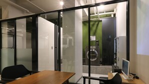 Un espace de bureau moderne avec une table en bois, des chaises noires, un ordinateur et une grande fenêtre donnant sur un mur végétal et un lave-linge.